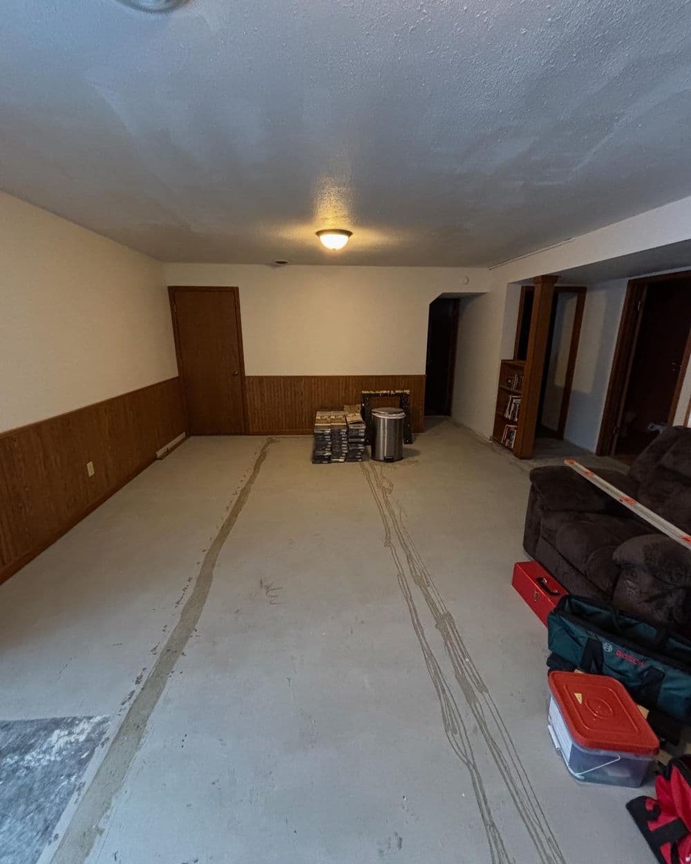 Empty living room with bare floor, wooden paneling, and renovation supplies stacked in the corner.
