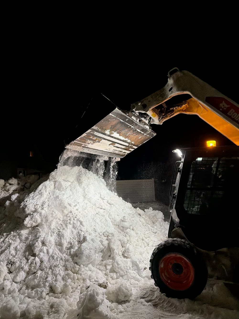 Skid steer loader dumping snow at night, creating a large snow pile.