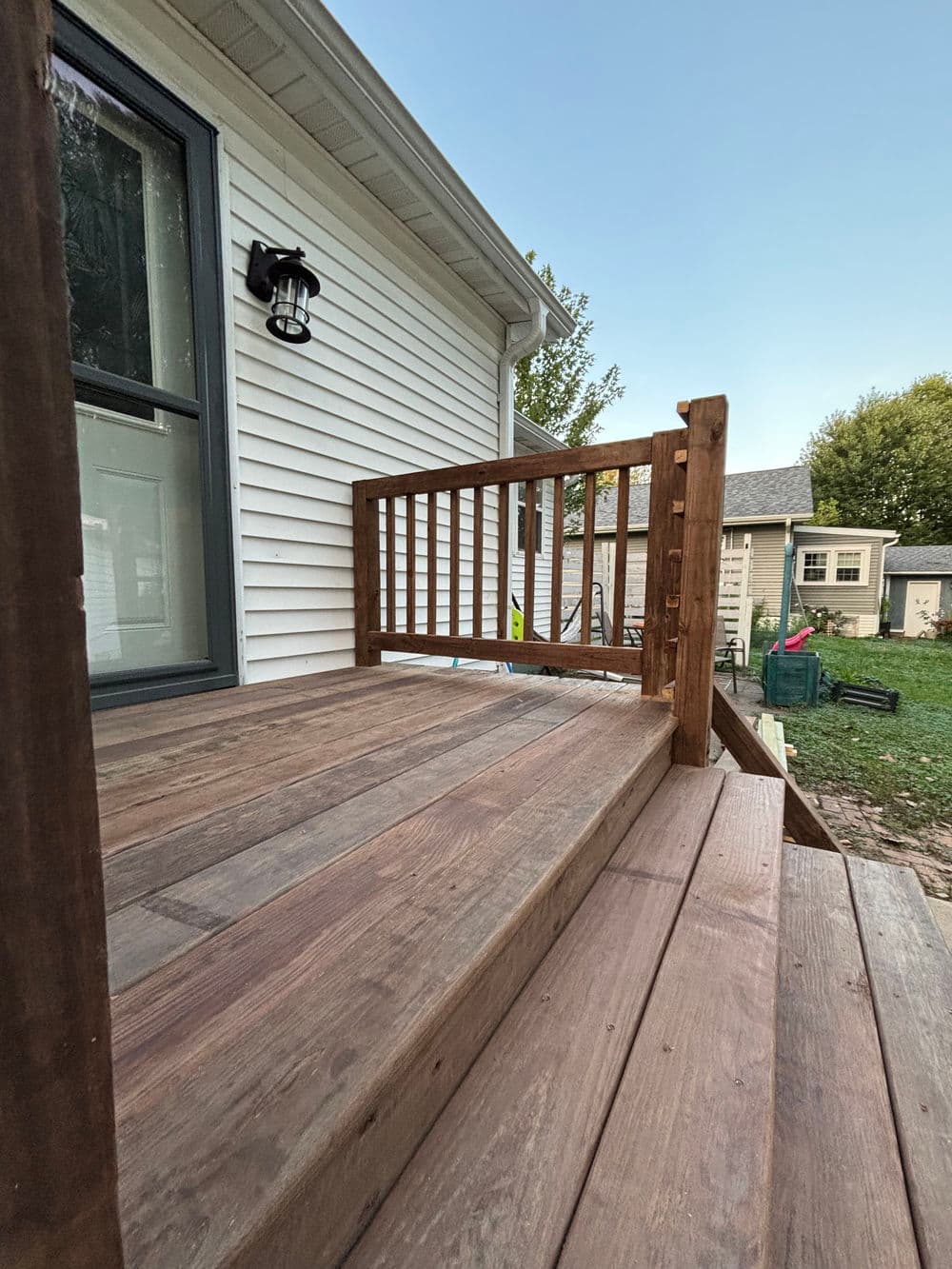 Wooden porch with railing and lantern, leading to grassy backyard and adjacent houses.