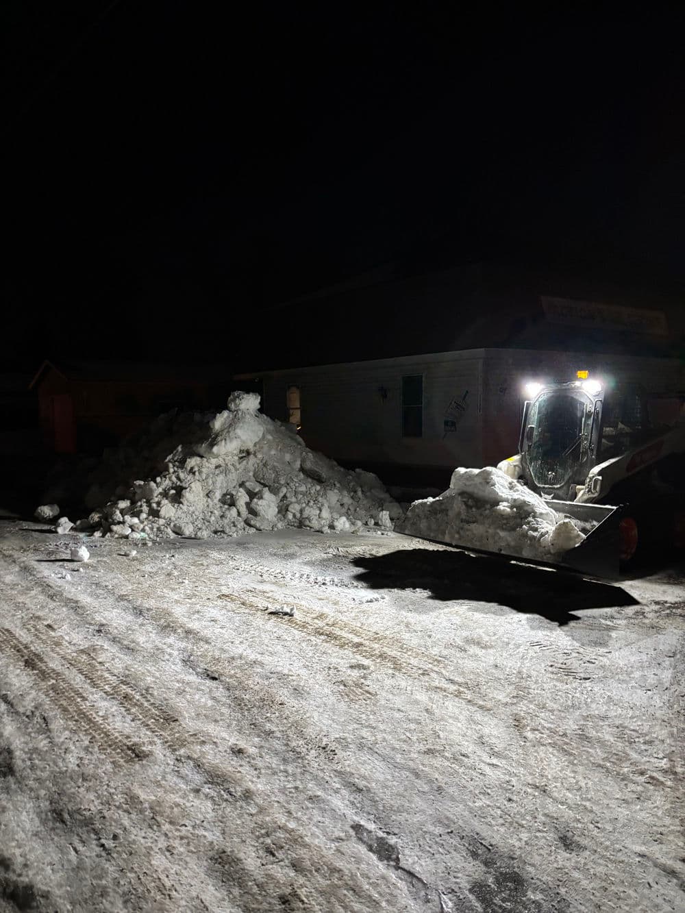 Snow removal at night near a building with a snow pile and a loader clearing the road.