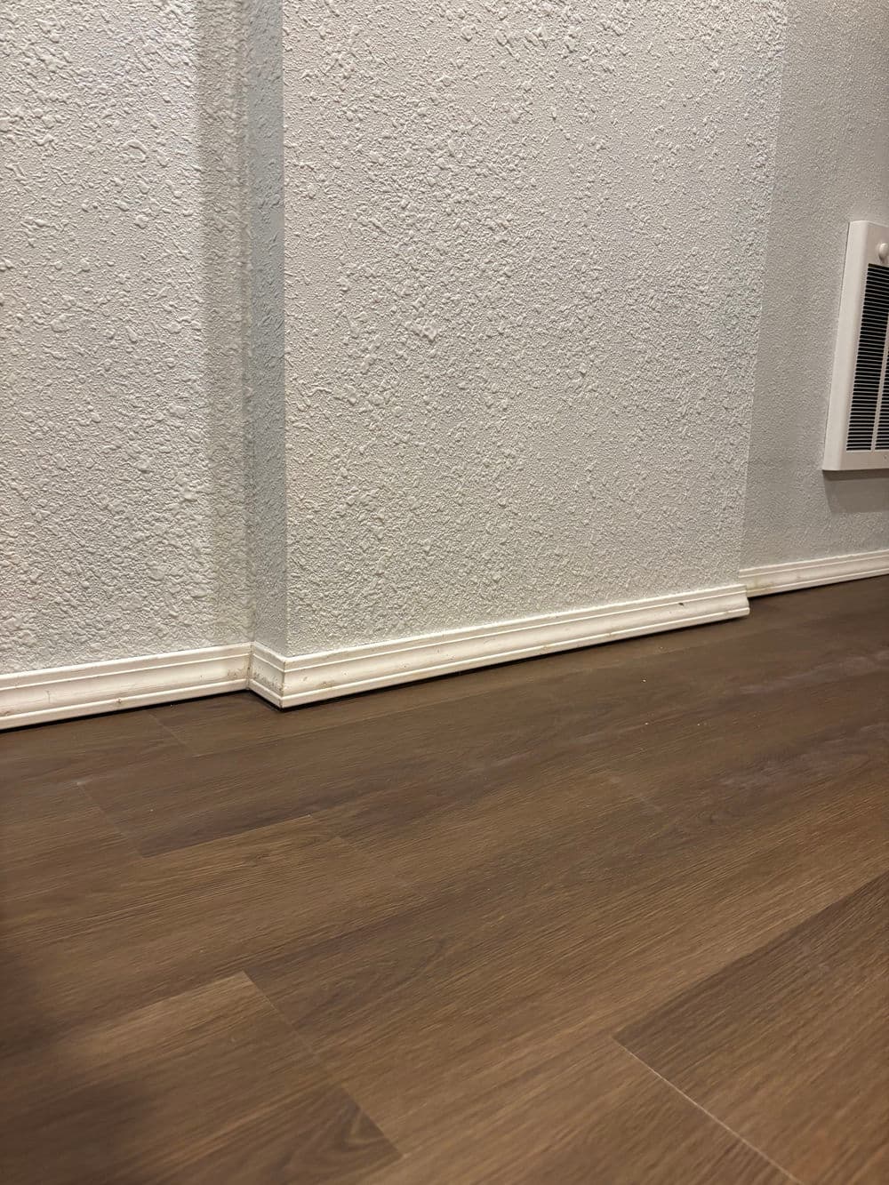 Interior corner with textured wall and wooden flooring, featuring white baseboard trim.