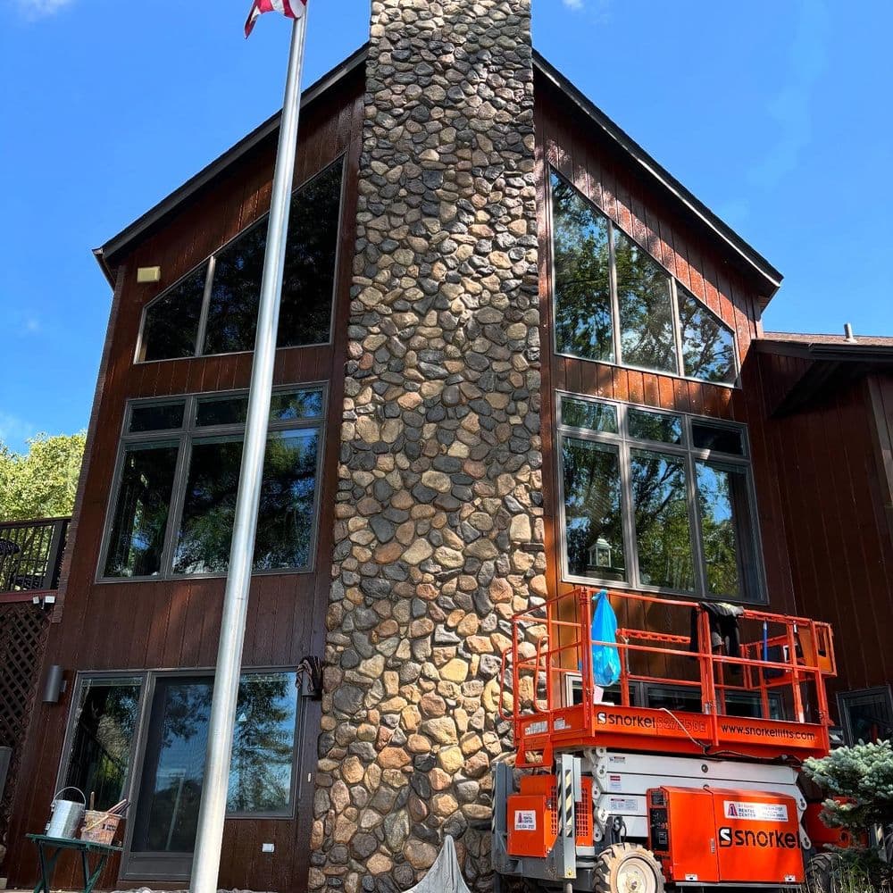 Modern wooden house with a tall stone chimney and construction lift, surrounded by trees.