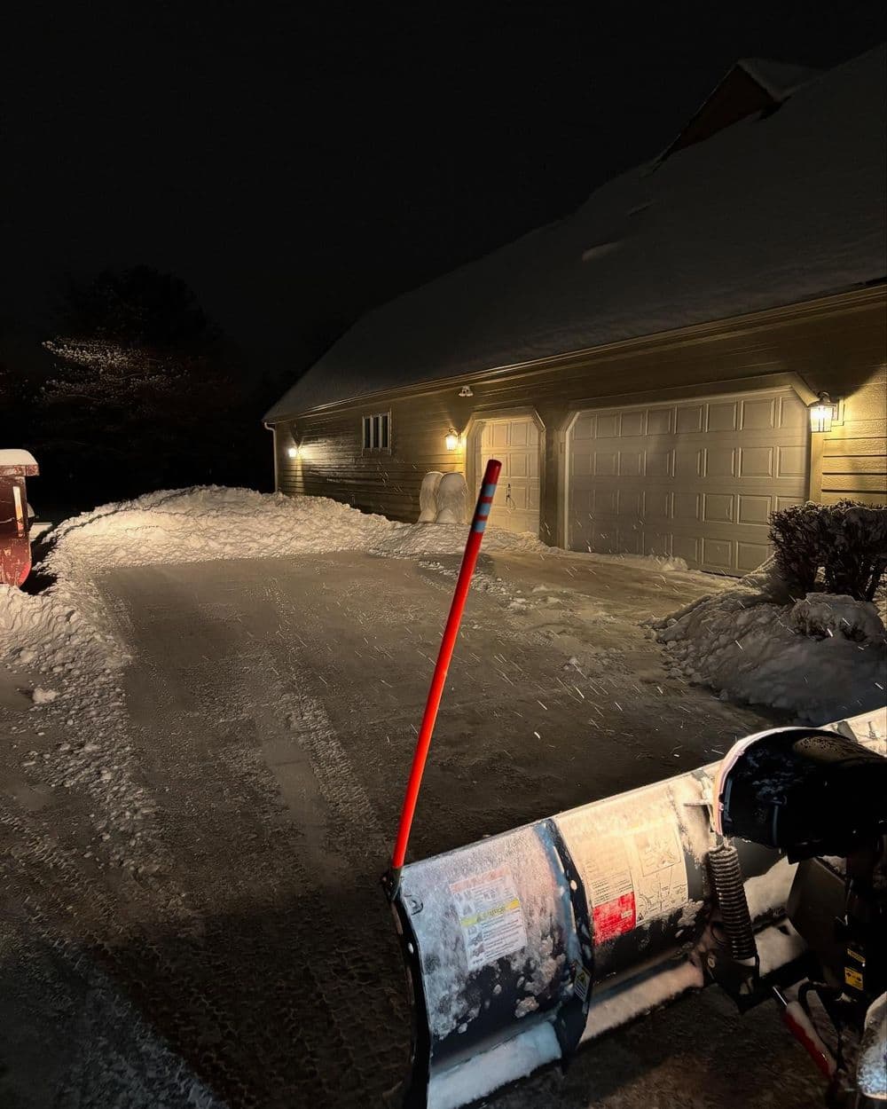 Snow-covered driveway at night with a snowplow in the foreground and a garage in the background.